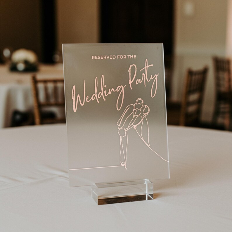 May include: A clear acrylic wedding sign with the text "RESERVED FOR THE Wedding Party" in a delicate pink script. A line drawing of a couple kissing is below the text. The sign is displayed on a clear acrylic base.