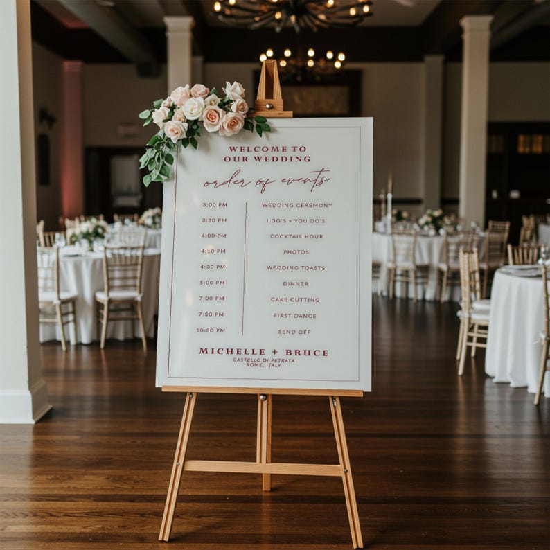 May include: A white sign with a burgundy border and text listing the order of events for a wedding. The sign is on an easel and has a floral arrangement on top. The text includes the wedding ceremony, cocktail hour, dinner, cake cutting, first dance, and send off. The sign also includes the names Michelle and Bruce and the location Castello di Petrata, Rome, Italy.