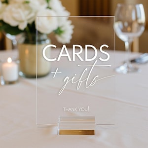 May include: A clear acrylic sign with white text that reads "CARDS + gifts THANK YOU!" The sign is on a gold stand and is sitting on a white tablecloth.