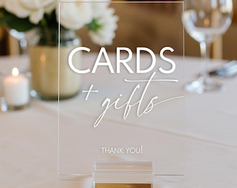 May include: A clear acrylic sign with white text that reads "CARDS + gifts THANK YOU!" The sign is on a gold stand and is sitting on a white tablecloth.