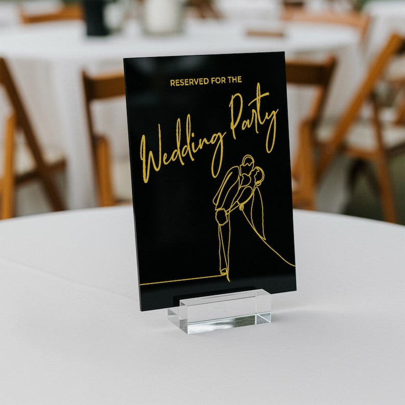 May include: A black acrylic sign with gold lettering and a line drawing of a couple. The sign reads "RESERVED FOR THE Wedding Party". The sign is displayed on a clear acrylic base, perfect for wedding decor.