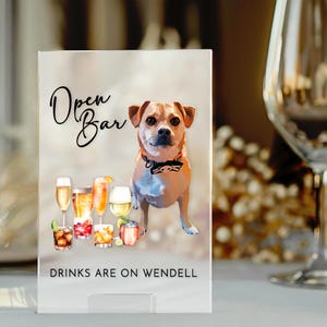 May include: A white acrylic sign with the text "Open Bar" in black cursive font. A cartoon image of a brown and white dog with a black bow tie is in the center of the sign. The text "Drinks are on Wendell" is at the bottom of the sign.