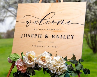 May include: Wooden wedding welcome sign on an easel. The sign reads "Welcome to our wedding Joseph & Bailey February 14, 2025" in black script. The sign is decorated with white and pink flowers and greenery.