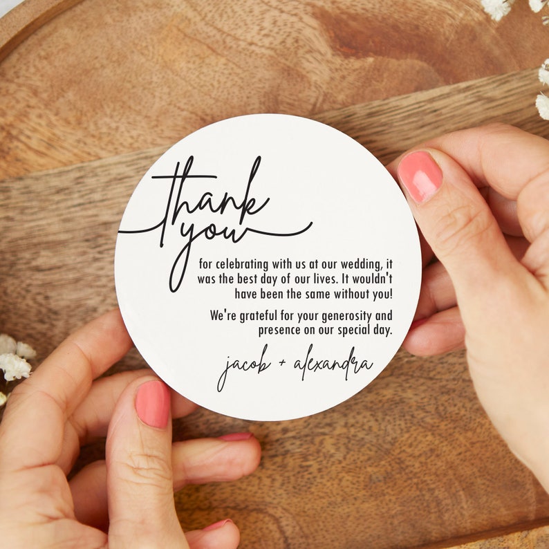 May include: White round thank you card with black calligraphy text that reads "Thank you for celebrating with us at our wedding, it was the best day of our lives. It wouldn't have been the same without you! We're grateful for your generosity and presence on our special day. jacob + alexandra"