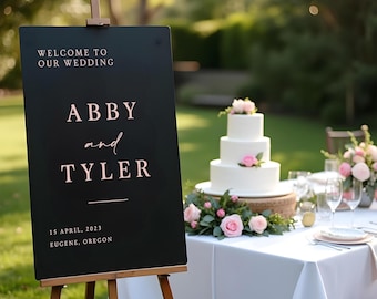 Puede incluir: Señal de boda negra en un caballete con el texto "Bienvenido a nuestra boda, Abby y Tyler, 15 de abril de 2023, Eugene, Oregón". Un pastel de boda blanco con flores rosas está en el fondo.
