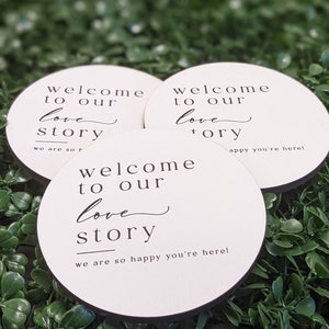 May include: Three round white signs with black text that reads "Welcome to our love story" and "We are so happy you're here!"