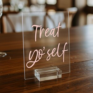 K&ouml;nnte beinhalten: Ein klares Acrylschild mit einer pinkfarbenen handgeschriebenen Nachricht, die "Treat yo' self" lautet. Das Schild steht auf einem klaren Acrylst&auml;nder.