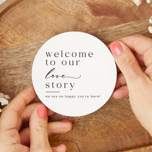May include: White round sign with black text that reads "Welcome to our love story" with a line under "story" and the text "we are so happy you're here!"
