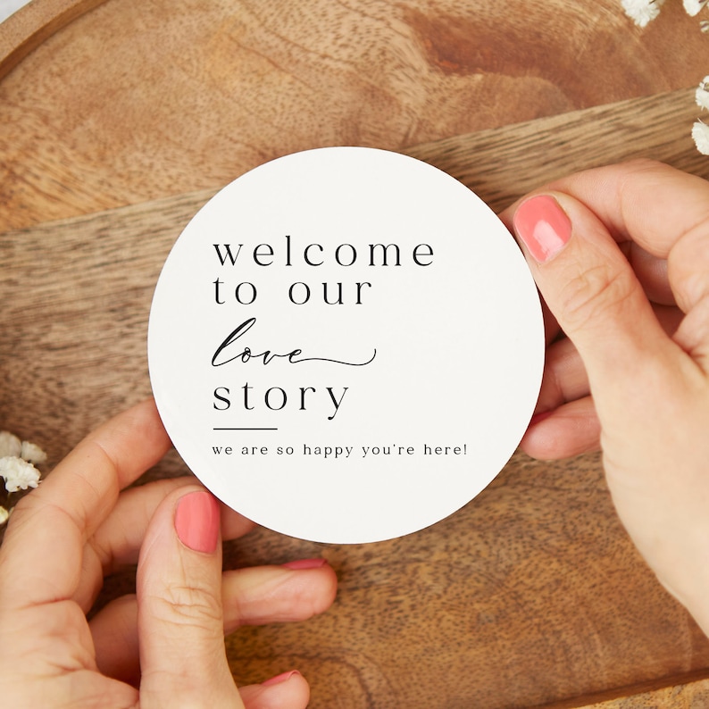 May include: White round sign with black text that reads "Welcome to our love story" with a line under "story" and the text "we are so happy you're here!"
