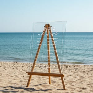 May include: A clear acrylic wedding sign with a white frame and white text. The sign lists the order of events for a wedding, including the wedding ceremony, cocktail hour, dinner, and first dance. The sign also includes the names "Michelle & Bruce" and the location "Castello di Prata, Italy".