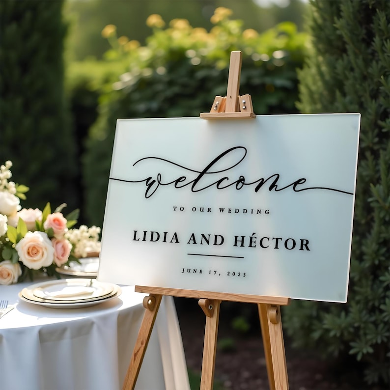May include: White frosted welcome sign with black calligraphy lettering on an easel. The sign reads "Welcome to our wedding Lidia and Héctor June 17, 2023".