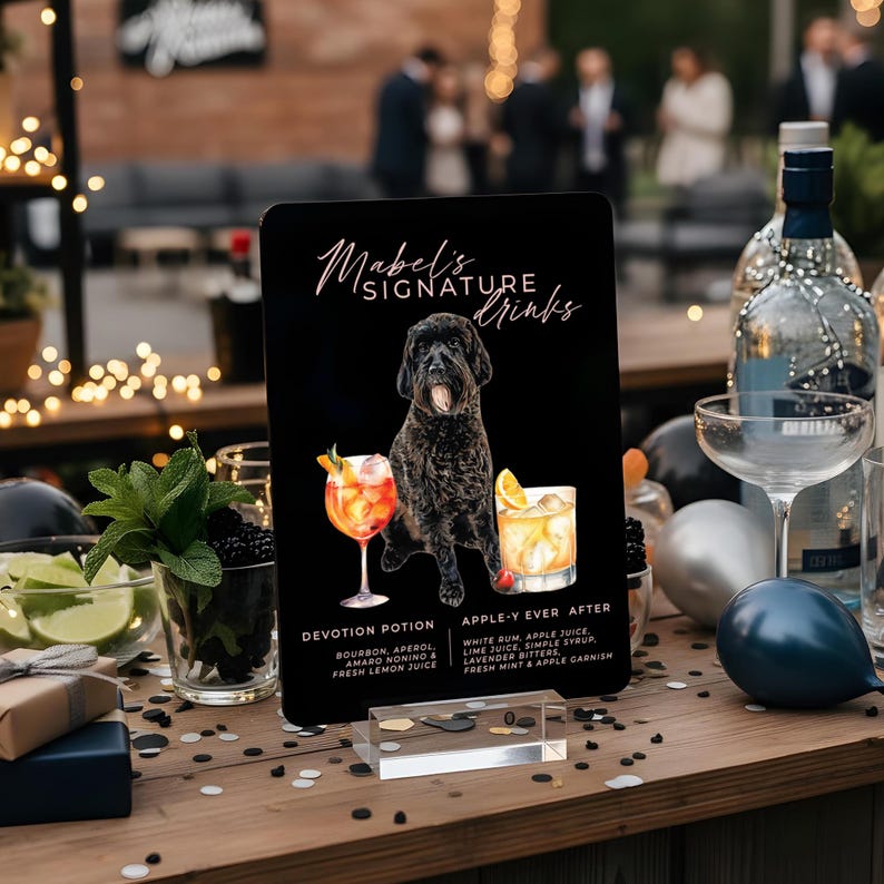May include: A black sign with the text "Mabel's Signature Drinks" and illustrations of two cocktails and a black dog. The sign lists the ingredients for each drink: Devotion Potion and Apple-Y Ever After. The sign is on a wooden table.