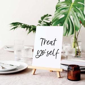 K&ouml;nnte beinhalten: Ein wei&szlig;es Schild mit schwarzer Kalligrafie, auf dem "Treat yo'self" steht, auf einem kleinen Holzstaffelei. Das Schild steht auf einem Tisch mit einer wei&szlig;en Tischdecke, klaren Gl&auml;sern und einem braunen Glasgef&auml;&szlig;.