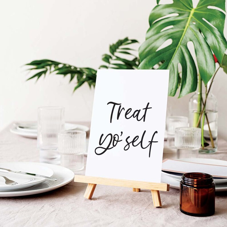 K&ouml;nnte beinhalten: Ein wei&szlig;es Schild mit schwarzer Kalligrafie, auf dem "Treat yo'self" steht, auf einem kleinen Holzstaffelei. Das Schild steht auf einem Tisch mit einer wei&szlig;en Tischdecke, klaren Gl&auml;sern und einem braunen Glasgef&auml;&szlig;.
