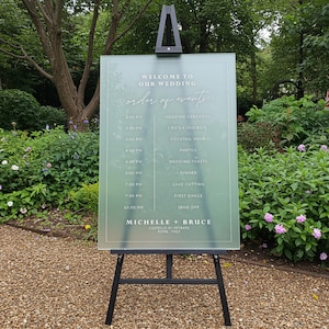 May include: A frosted acrylic wedding sign with a light blue background and white text. The sign lists the order of events for a wedding, including the wedding ceremony, cocktail hour, dinner, and first dance. The sign also includes the names of the bride and groom, Michelle and Bruce, and the location of the wedding, Castello di Petrata, Rome, Italy.