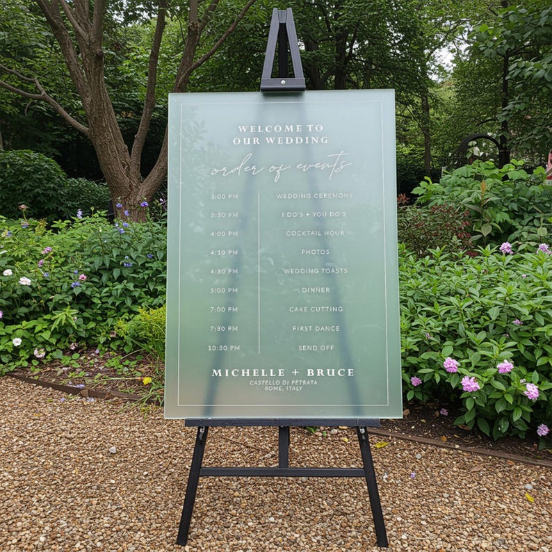 May include: A frosted acrylic wedding sign with a light blue background and white text. The sign lists the order of events for a wedding, including the wedding ceremony, cocktail hour, dinner, and first dance. The sign also includes the names of the bride and groom, Michelle and Bruce, and the location of the wedding, Castello di Petrata, Rome, Italy.