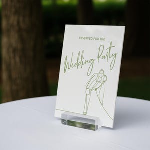 May include: A white sign with the text "RESERVED FOR THE Wedding Party" in green script. A line drawing of a couple kissing is below the text. The sign is held by a clear acrylic stand, placed on a white surface.