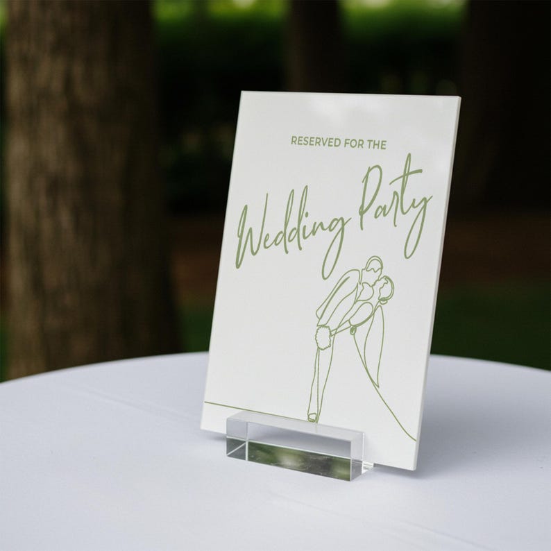 May include: A white sign with the text "RESERVED FOR THE Wedding Party" in green script. A line drawing of a couple kissing is below the text. The sign is held by a clear acrylic stand, placed on a white surface.