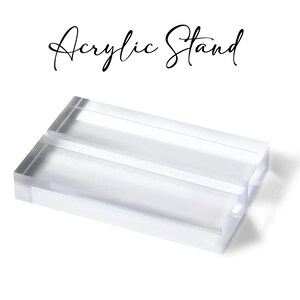 May include: Clear acrylic stand with a rectangular shape and a slot in the center. The words "Acrylic Stand" are written in a cursive font above the stand. The stand is on a white background.