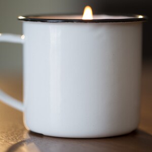May include: A white enamel mug with a black rim, filled with a white candle that is lit.