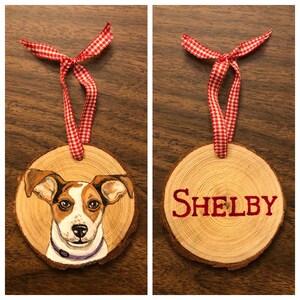 May include: A wooden Christmas ornament with a painted portrait of a Jack Russell Terrier dog. The ornament is decorated with a red and white gingham ribbon and the name "SHELBY" is painted on the back.