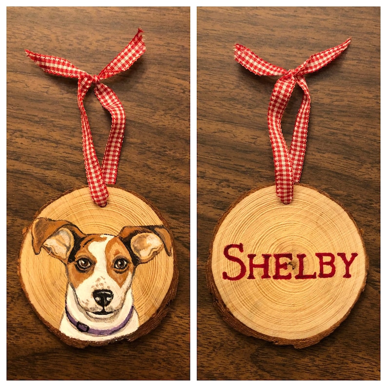 May include: A wooden Christmas ornament with a painted portrait of a Jack Russell Terrier dog. The ornament is decorated with a red and white gingham ribbon and the name "SHELBY" is painted on the back.