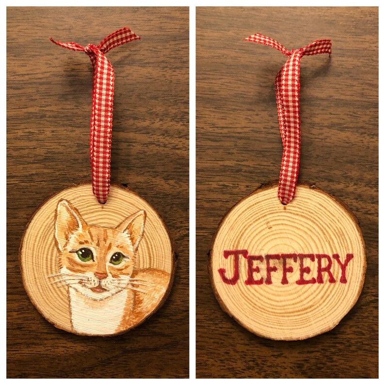 May include: Two wooden ornaments with red and white gingham ribbon. One ornament features a hand-painted portrait of a ginger cat. The other ornament has the name "JEFFERY" painted in red.