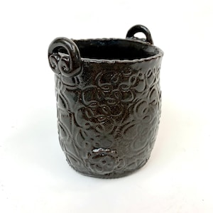 May include: A dark brown ceramic planter with two loop handles. The planter has a textured surface with a floral design.