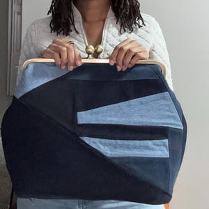 May include: A large clutch bag featuring a patchwork design of dark navy and light blue denim fabric panels. The bag has a light-colored wooden frame and a metallic kiss-lock closure with two round brass-toned knobs at the top center.