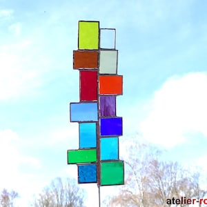 Könnte beinhalten: Ein farbenfrohes Mobile aus Glasmosaik mit Quadraten aus gelbem, orangefarbenem, rotem, blauem, grünem und violettem Glas. Das Mobile hängt an einer Metallstange.