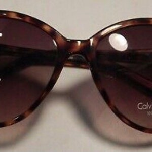 May include: Tortoise shell sunglasses with brown lenses and a pink accent on the frame. The sunglasses have the text "Calvin Klein Jeans 100% UV protection" printed on the right lens.
