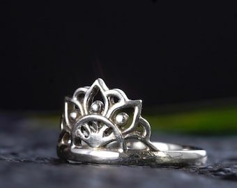 Könnte beinhalten: Ein silberner Ring mit einem filigranen, kunstvollen Design, das an eine stilisierte Blume oder ein Mandala erinnert. Der Ring ist vor einem dunklen Hintergrund platziert.