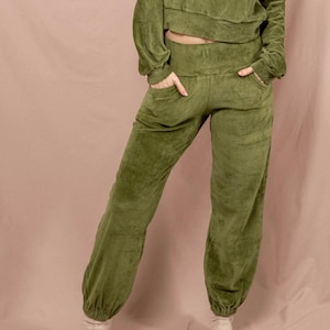 Könnte beinhalten: Olivgrünes Velours-Loungewear-Set. Das Set besteht aus einem cropped Oberteil mit langen Ärmeln und einer passenden hoch taillierten Hose mit Taschen. Die Hose hat eine lockere Passform mit elastischen Bündchen an den Knöcheln. Die Person trägt weiße Socken.