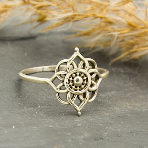 Könnte beinhalten: Ein silberfarbener Ring mit floralem Design. Der Ring zeigt eine filigrane Blume mit einem erhabenen Mittelkreis und blütenblattförmigen Elementen. Er liegt auf einer dunkelgrauen Oberfläche, mit getrocknetem Gras im Hintergrund.