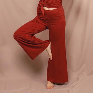 Può includere: Un completo da yoga color ruggine, composto da un top a maniche lunghe avvolgente e pantaloni a gamba larga. L'outfit è realizzato in un tessuto morbido e fluido. La modella è in posa yoga con le mani giunte in preghiera.