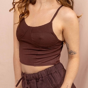 Könnte beinhalten: Ein zweiteiliges Outfit in Braun. Das Set besteht aus einem gerippten Crop-Top mit dünnen Trägern und einer passenden Hose mit hohem Bund. Die Hose hat einen Kordelzug in der Taille und ein dezentes Fischgrätenmuster. Das Outfit ist aus weichem Material.