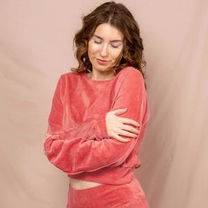 Könnte beinhalten: Ein korallenfarbenes Velours-Loungewear-Set, bestehend aus einem gecroppten Sweatshirt mit Rundhalsausschnitt und einer hoch taillierten Hose. Der weiche, strukturierte Stoff schafft ein komfortables und stilvolles Outfit.