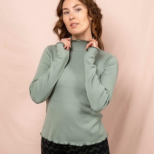 Könnte beinhalten: Ein salbeigrünes Langarm-Top mit hohem, gerüschtem Ausschnitt und Manschetten. Das Top wird mit einer schwarzen gemusterten Leggings kombiniert. Das Model trägt das Top und die Leggings vor einem rosa Hintergrund. Das Top hat eine gerippte Textur.