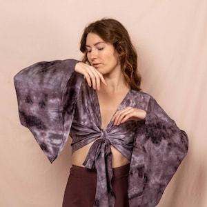 Op de afbeelding: Een crop top met strik en wijde, klokmouwen. De top heeft een donkerpaars tie-dye patroon. Het model draagt een bijpassende donkerbruine rok. De top heeft een diepe V-hals.