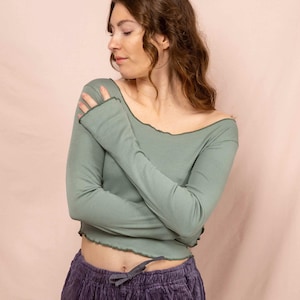 Könnte beinhalten: Salbeigrünes Langarm-Top mit gewelltem Ausschnitt und Manschetten. Das Top wird mit einer lila Cordhose mit Kordelzug getragen. Das Outfit ist lässig und bequem, geeignet für den Alltag.