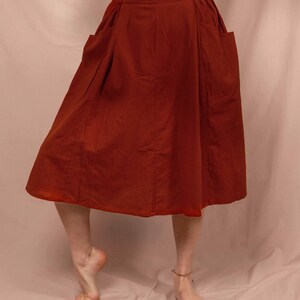 Könnte beinhalten: Rostfarbener Midirock mit zwei großen Vordertaschen. Der Rock hat eine hohe Taille und eine ausgestellte Silhouette. Der Stoff scheint mittelschweres Baumwoll- oder Leinenmaterial zu sein. Das Model trägt ein langärmliges, neutralfarbenes Oberteil.