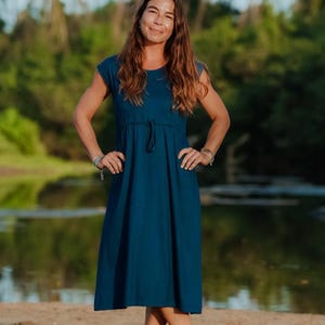 Könnte beinhalten: Ein teal-farbenes Kleid mit kurzen Ärmeln und einem Kordelzug in der Taille. Das knielange Kleid wird von einer Person getragen, die an einem sandigen Strand steht. Der Hintergrund zeigt ein Gewässer und üppiges grünes Laub.