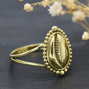 Könnte beinhalten: Ein goldfarbener Ring mit einem detaillierten Kaurischalen-Design. Der Ring hat einen perlenbesetzten Rand und ein schlichtes Band.