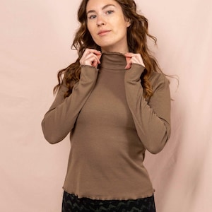 Könnte beinhalten: Ein langärmliges, braunes Rollkragenshirt mit Rippstruktur. Das Shirt hat einen hohen Ausschnitt und eine figurbetonte Silhouette. Das Model trägt eine Hose mit schwarzem und grünem Muster. Der Hintergrund ist einfarbig hellrosa.