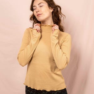 Könnte beinhalten: Senfgelbes Langarm-Top mit gerippter Struktur und Rüschenausschnitt und -saum. Das Model trägt eine schwarz-grüne gemusterte Leggings. Das Top hat einen hohen, gerüschten Ausschnitt.