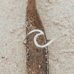Könnte beinhalten: Ein silberner Ring in Wellenform, der auf einem Stück verwittertem Holz vor einem sandigen Hintergrund liegt. Das Design des Rings ist eine einfache, elegante Welle, die die Essenz des Ozeans einfängt. Der Ring ist ein Schmuckstück.