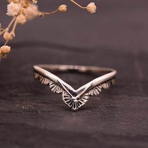 Peut inclure: Une bague argentée en forme de V. La bague présente un motif décoratif détaillé sur les bords et au centre. La bague est présentée sur une surface sombre et texturée, avec des fleurs séchées en arrière-plan.