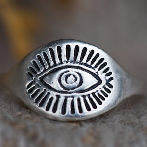 Könnte beinhalten: Ein silberner Ring mit einem Augenmotiv. Das Auge ist von einem Kreis aus Strahlen umgeben.