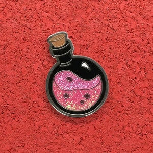 Könnte beinhalten: Eine schwarze und silberne Emaille-Anstecknadel mit einer Zaubertrankflasche mit pinkfarbener Glitzerflüssigkeit im Inneren. Die Flasche hat einen Korkverschluss.
