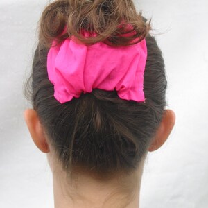 May include: A woman with brown hair is wearing a bright pink scrunchie to hold her hair up in a bun.